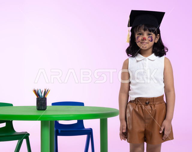 التلوين والرسم بألوان صحية على وجوه الأطفال، رسومات وجه للبنات تدل على الأنوثة واللطافة، بورتريه لطفلة عربية خليجية سعودية ترتدي الزي الكاجوال وقبعة التخرج من الروضة، إيماءات وجه تدل على الحزن والرغبة بالبكاء، خلفية وردية.