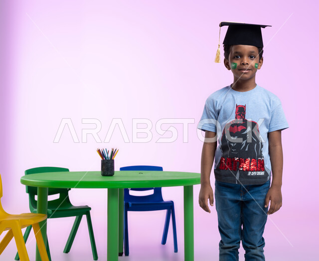 إيماءات تدل على الثقة بالنفس، التلوين والرسم بألوان صحية على وجوه الأطفال، أشكال ورسومات وجه للأولاد تدل على اللطافة، بورتريه لطفل عربي خليجي سعودي يرتدي الزي الكاجوال وقبعة التخرج من الروضة، خلفية وردية.