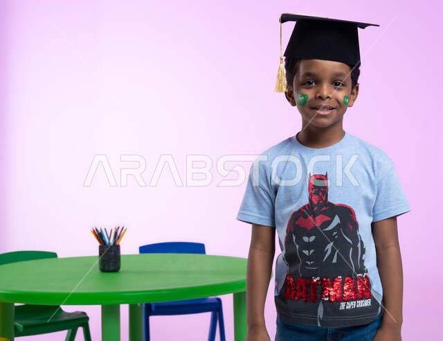 التلوين والرسم بألوان صحية على وجوه الأطفال، أشكال ورسومات وجه للأولاد تدل على اللطافة، بورتريه لطفل عربي خليجي سعودي يرتدي الزي الكاجوال وقبعة التخرج من الروضة، إيماءات تدل على الثقة بالنفس، خلفية وردية.