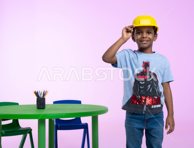 دمج التعليم باللعب في الروضة، تعريف الأطفال على مفهوم الهندسة والمهن المستقبلية، بورتريه مقرب لفتى عربي خليجي سعودي مبتسم، يرتدي خوذة الوقاية الخاصة بالعمل، ينظر إلى الكاميرا بإيماءات وجه ويدين مختلفة، مهنة ووظيفة هندسية، خلفية زهرية.
