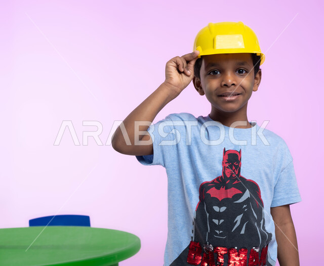 تعريف الأطفال على مفهوم الهندسة والمهن المستقبلية، بورتريه مقرب لفتى عربي خليجي سعودي مبتسم في الروضة، يرتدي خوذة الوقاية الخاصة بالعمل، ينظر إلى الكاميرا بإيماءات وجه ويدين مختلفة، مهن ووظائف هندسية، خلفية زهرية.