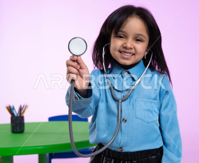 تعريف الأطفال على المهن المستقبلية، مفهوم الطب والرعاية الصحية، بورتريه مقرب لفتاة عربية خليجية سعودية في الروضة، تقوم بالفحص بواسطة السماعة الطبية، إيماءات وجه تدل على الفرح والسعادة، خلفية زهرية.