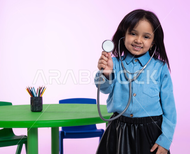  إيماءات وجه تدل على الفرح والسعادة، تعريف الأطفال على المهن المستقبلية، مفهوم الطب والرعاية الصحية، بورتريه مقرب لفتاة عربية خليجية سعودية في الروضة، تقوم بالفحص بواسطة السماعة الطبية، خلفية زهرية.