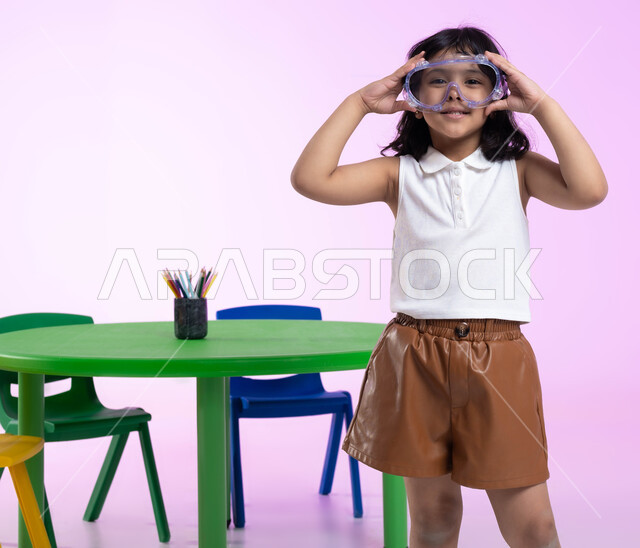 الحماس والشوق للغوص في الماء، الاستعداد والتحضير للسباحة، بورتريه لطفلة عربية خليجية سعودية ترتدي الزي الكاجوال ونظارات السباحة، اللعب واللهو بأدوات المسبح، خلفية وردية.