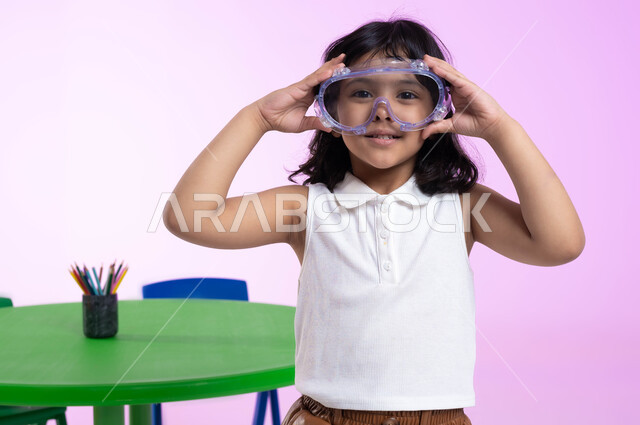 الاستعداد والتحضير للسباحة، بورتريه مقرب لطفلة عربية خليجية سعودية ترتدي الزي الكاجوال ونظارات السباحة، ايماءات الحماس والشوق للغوص في الماء، اللعب واللهو بأدوات المسبح، خلفية وردية.