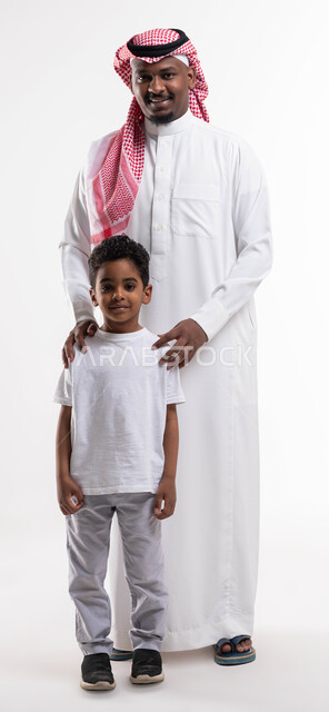 صورة الجسم بالطول الكامل، التشابه بين الاب و ابنه بالصفات و الجينات الوراثية، بورتريه مقرب لرجل عربي خليجي سعودي يرتدي الزي السعودي التقليدي برفقة ابنه بإيماءات تدل على السعادة، الاهتمام والاعتناء بالأبناء، مفهوم الأسرة السعيدة، خلفية بيضاء