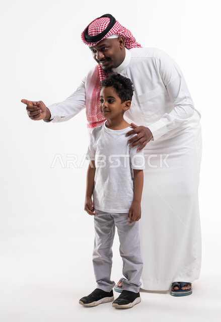 مفهوم تربية الابناء على الطريق الصحيح والاهتمام بهم، بورتريه لرجل عربي خليجي سعودي يرتدي  الثوب والشماغ السعودي برفقة ولده يشير بأصبعه للدلالة على شيء ما، لفت الانتباه لشيء معين،  صورة الجسم بالطول الكامل, خلفية بيضاء