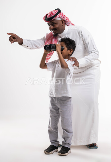 رؤية الأشياء البعيدة، تشجيع الأهل للأبناء على الاستكشاف والمغامرات ،بورتريه مقرب لولد عربي خليجي سعودي رفقة والده يحمل بيده منظار ثنائي العينين يقوم بالتخطيط المستقبلي للأهداف، الاشارة الى شيء ما، صورة الجسم بالطول الكامل، خلفية بيضاء