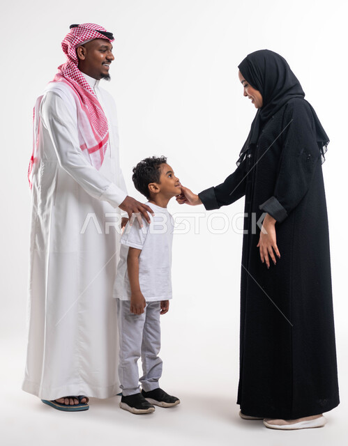 الإعتناء و الاهتمام بالأبناء، أجواء عائلية مليئة بالحب، بورتريه لسيدة عربية خليجية سعودية محجبة ترتدي العباءة الخليجية وتقوم بمداعبة ابنها برفقة زوجها بإيماءات السعادة ،مفهوم الاستقرار الأسري، الحياة الزوجية الناجحة، صورة الجسم بالطول الكامل، خلفية بيضاء