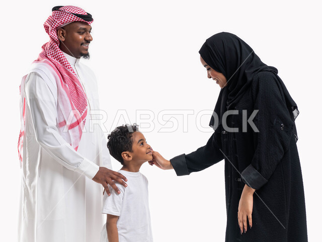 مفهوم الاستقرار الأسري، الإعتناء و الاهتمام بالأبناء، أجواء عائلية مليئة بالحب، بورتريه لسيدة عربية خليجية سعودية محجبة ترتدي العباءة الخليجية وتقوم بمداعبة ابنها برفقة زوجها بإيماءات السعادة , الحياة الزوجية الناجحة، خلفية بيضاء