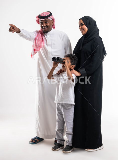  تشجيع الأهل للأبناء على الاستكشاف والمغامرات ،بورتريه لولد عربي خليجي سعودي رفقة والديه يحمل بيده منظار ثنائي العينين لرؤية الأشياء البعيدة، الاشارة الى شيء ما ، التخطيط المستقبلي للأهداف, ارتداء الزي التقليدي السعودي, صورة الجسم بالطول الكامل ،خلفية بيض