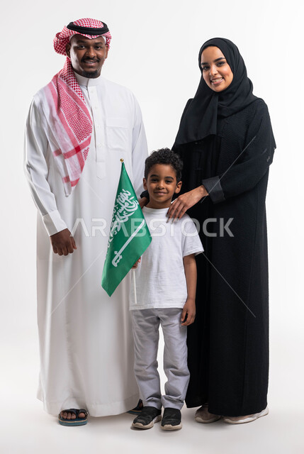 ذكرى يوم العلم السعودي ،بورتريه لولد عربي خليجي سعودي يحمل بيده علم المملكة العربية السعودية برفقة والديه بإيماءات تدل على السعادة، الإحتفال باليوم الوطني السعودي، راية التوحيد والإسلام، صورة الجسم بالطول الكامل، خلفية بيضاء