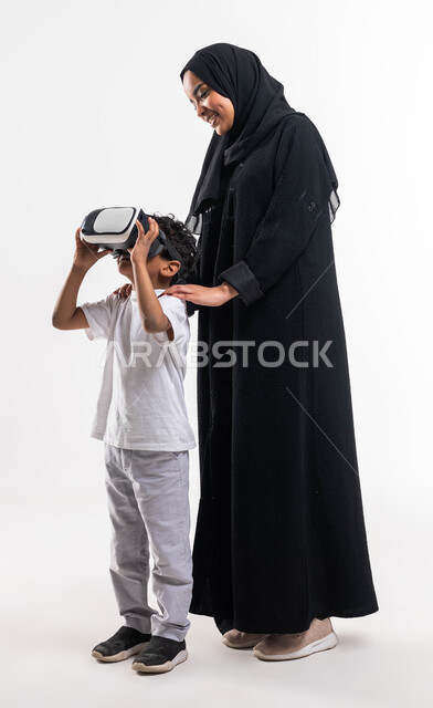 أنشطة ترفيهيه تقنية مسلية، بورتريه لشابة عربية خليجية سعودية محجبة ترتدي العباءة السعودية وتقف بجانب ابنها وهو يرتدي نظارة الواقع الإفتراضي ثلاثية الأبعاد، قضاء أوقات ممتعة مع الأبناء بمشاهدة الأفلام والمسلسلات، صورة الجسم بالطول الكامل، خلفية بيضاء