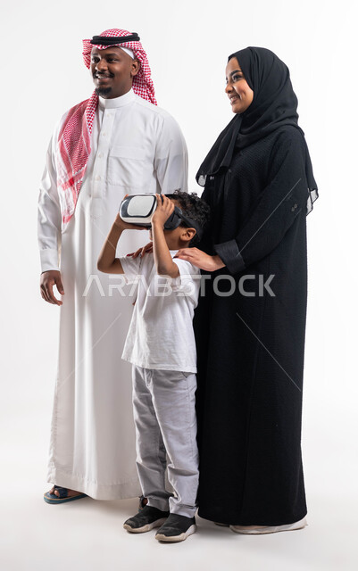 قضاء أوقات ممتعة  مع الأبناء بمشاهدة الأفلام والمسلسلات، أنشطة تقنية ترفيهية مسلية، بورتريه لعائلة عربية خليجية سعودية مكونة من الأب و الأم و الابن بإيماءات السعادة ،ارتداء نظارة الواقع الافتراضي ثلاثية الأبعاد, صورة الجسم بالطول الكامل, خلفية بيضاء