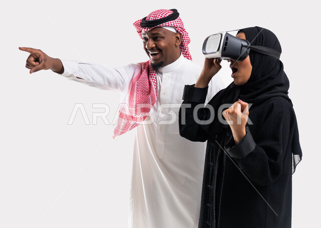 قضاء أوقات ممتعة بمشاهدة مباريات كرة القدم, أنشطة تقنية ترفيهية مسلية, الاستمتاع بمشاهدة الأفلام، بورتريه لرجل عربي خليجي سعودي يشير بأصبعه الى شيء ما يقف بجانب زوجته وهي ترتدي نظارة الواقع الافتراضي, ايماءات السعادة بالفوز والانتصار، خلفية بيضاء