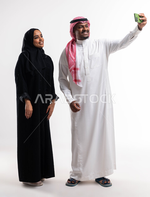 التقاط صور السيلفي عبر الجوال للذكريات العائلية الودية، بورتريه لرجل عربي خليجي سعودي يرتدي الثوب والشماغ السعودي برفقة زوجته، قضاء أوقات ممتعه مع الاهل، توثيق اللحظات الأسرية السعيدة، ايماءات تدل على السعادة، صورة الجسم بالطول الكامل، خلفية بيضاء