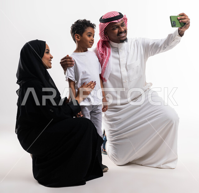 التقاط صور السيلفي عبر الجوال للذكريات العائلية الودية، الاهتمام والاعتناء بالأبناء، بورتريه لسيدة عربية خليجية سعودية محجبة برفقة ابنها وزوجها ، قضاء أوقات ممتعه مع الاهل، توثيق اللحظات الأسرية السعيدة, ايماءات تدل على السعادة، خلفية بيضاء