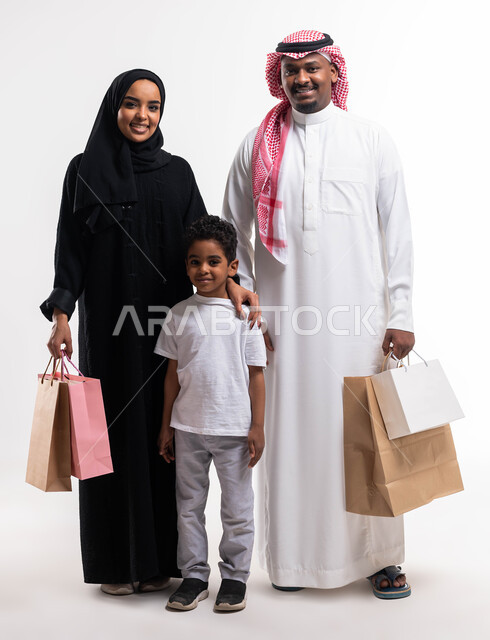 ايماءات تدل على السعادة، تلبية الاحتياجات المنزلية، متعة الشراء في السعودية، بورتريه لعائلة عربية خليجية سعودية تحمل حقائب التسوق رفقة ابنهما ، أساسيات العائلة السعوديه السعيده، الاستفادة من العروض والتنزيلات, صورة الجسم بالطول الكامل، خلفية بيضاء