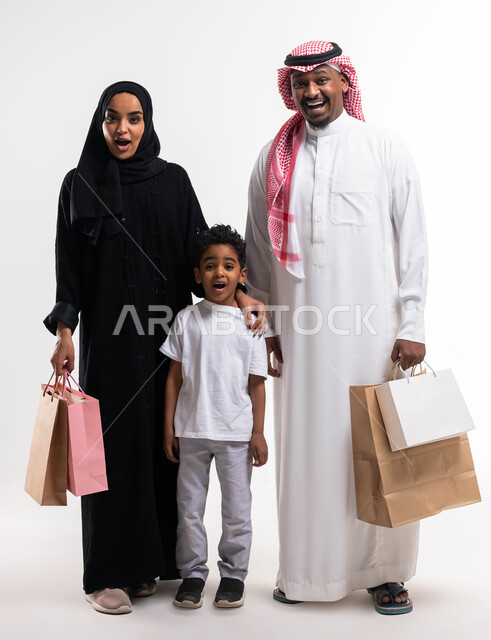ايماءات تدل على الدهشة والذهول بالأسعار، متعة الشراء في السعودية، بورتريه لعائلة عربية خليجية سعودية تحمل حقائب التسوق رفقة ابنهما في المول، أساسيات العائلة السعوديه السعيده، الاستفادة من العروضات والخصومات, صورة الجسم بالطول الكامل، خلفية بيضاء