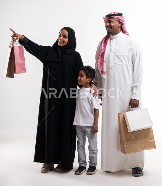 الاشارة الى شيء ما بإيماءات السعادة، متعة الشراء في السعودية، بورتريه لزوجان عربيان خليجيان سعوديان يحملان حقائب التسوق رفقة ابنهما في المول، أساسيات العائلة السعوديه السعيده، الحياة الزوجية الناجحة، صورة الجسم بالطول الكامل، خلفية بيضاء