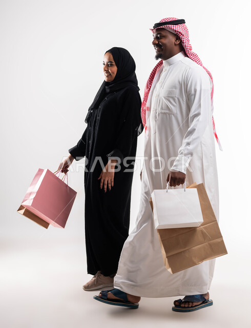 الاستفادة من العروض والخصومات، تلبية الاحتياجات المنزليه، بورتريه لزوجان عربيان خليجيان سعوديان يحملان حقائب التسوق، ايماءات تدل على السعادة، أساسيات العائلة السعوديه السعيده، الحياة الزوجية الناجحة, صورة الجسم بالطول الكامل، خلفية بيضاء
