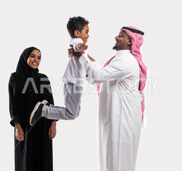 مفهوم الاهتمام والاعتناء بالأطفال, اللعب والتسلية واالترفيه ،بورتريه لعائلة عربية خليجية سعودية تقضي أوقات ممتعه مع الأبناء، رجل سعودي يرفع ابنه عاليا بإيماءات تدل على السعادة، المحافظة على ترابط الأسرة، خلفية بيضاء