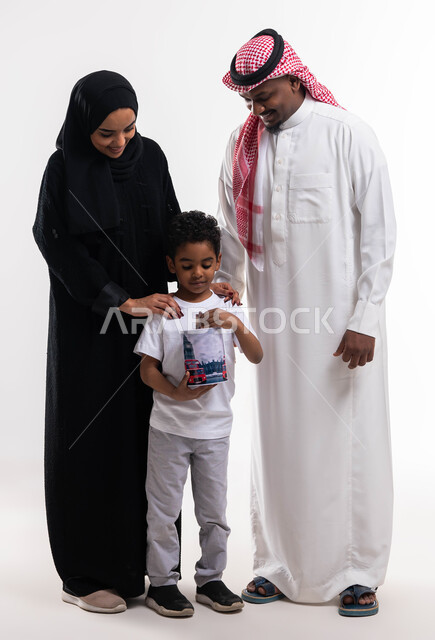 تعليم الأطفال التوفير و تجميع المال ،بورتريه لوالدان عربيان خليجيان سعوديان مع ابنهما يجلسان على كنبة رمادية في غرفة المعيشة ، تجميع المصروف داخل صندوق المال ( الحصالة ) ،زيادة وعي الطفل ،صورة الجسم بالطول الكامل، خلفية بيضاء