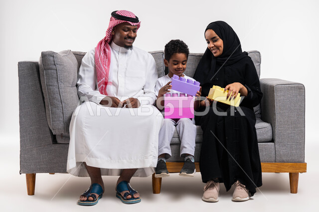 انشطة ترفيهية مفيدة، تعزيز المهارات الفكرية، بورتريه لعائلة عربية سعودية  تجلس على كنبة رمادية في غرفة المعيشة، ايماءات وجه تدل على السعادة، تنمية القدرات على الابتكار، قضاء أوقات ممتعه مع الاهل والأبناء، خلفية بيضاء