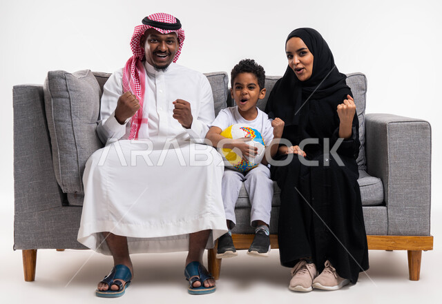 ايماءات  السعادة بالانتصار والفوز، أندية وتشجييع, بورتريه لعائلة عربية خليجية سعودية تجلس على كنبه رماديه في غرفة المعيشة و تشاهد مباريات كرة القدم ، قضاء الوقت مع الأهل والأبناء ، الشغف في متابعه فريق كرة القدم ،خلفية بيضاء