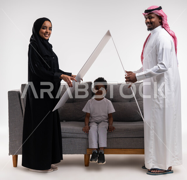 بناء مجتمع سعودي متنامي، اسكان وتمليك ،الجلوس على كنبه رماديه في غرفة المعيشة ,بورتريه لزوجان عربيان سعوديان خليجيان يحملان لوح أبيض فوق رأس الابن، قضاء الوقت مع الأهل والأبناء ،تلبية الاحتياجات المنزليه العائلية، ايماءات تدل على السعادة، خلفية بيضاء .