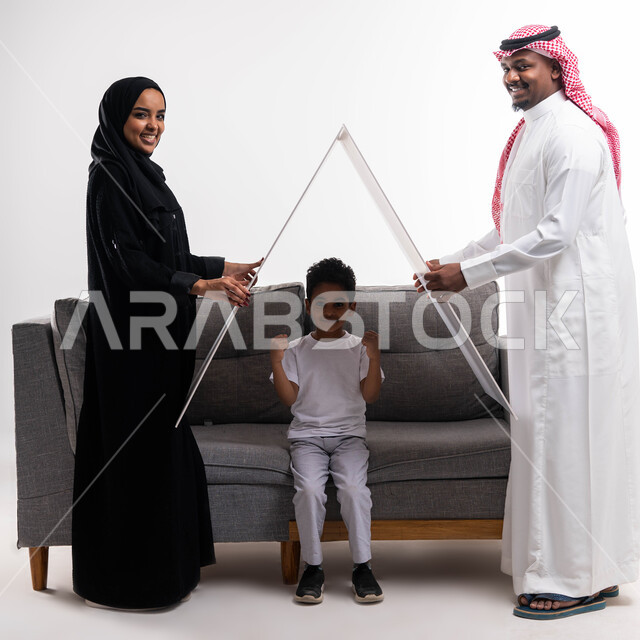 تلبية الاحتياجات المنزليه العائلية، ايماءات تدل على السعادة, الاستقرار العائلي في الاسكان والتمليك ،بورتريه لزوجان عربيان سعوديان خليجيان  يحملان لوح أبيض فوق رأس الابن وهو يجلس على كنبه رماديه في غرفة المعيشة , قضاء الوقت مع الأهل والأبناء ، خلفية بيضاء