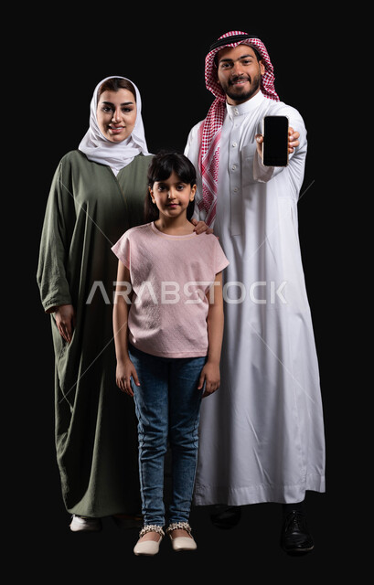 التسويق الإلكتروني وعرض خدمات وتطبيقات الجوال، بورتريه لرجل عربي سعودي خليجي مع عائلته يمسك الهاتف المحمول بشاشة سوداء فارغة، الإهتمام والإعتناء بالعائلة، المحافظة على الترابط الأسري والإستقرار العائلي، صورة الجسم بالطول الكامل، خلفية سوداء