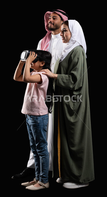 أنشطة تقنية ترفيهية مسلية، بورتريه لفتاة عربية سعودية خليجية برفقة والديها ترتدي نظارات الواقع الإفتراضي (في آر) ثلاثية الإبعاد، مفهوم الإهتمام والإعتناء بالأبناء، الإستمتاع بمشاهدة الأفلام ومحاكاة العالم الإفتراضي، صورة الجسم بالطول الكامل، خلفية سوداء