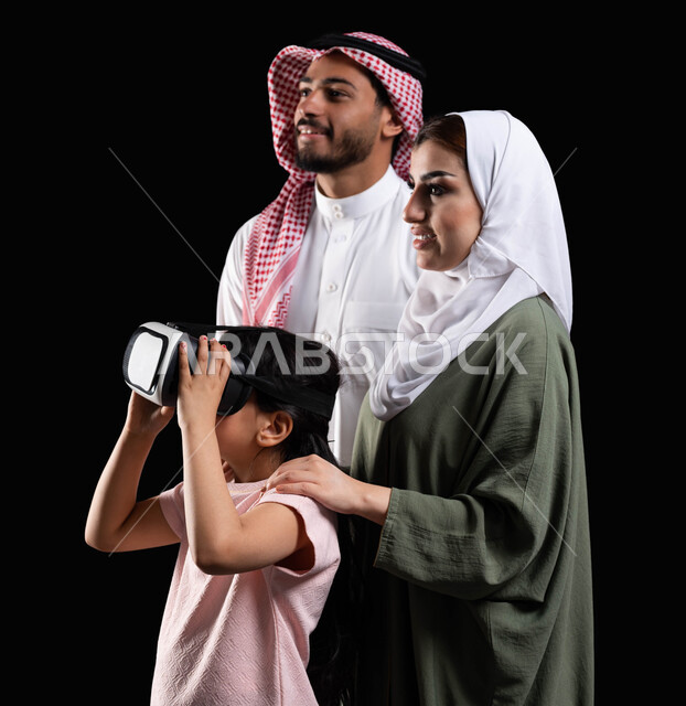 مفهوم الإهتمام والإعتناء بالأبناء، أنشطة تقنية ترفيهية مسلية، بورتريه لفتاة عربية سعودية خليجية برفقة والديها ترتدي نظارات الواقع الإفتراضي (في آر) ثلاثية الإبعاد، الإستمتاع بمشاهدة الأفلام ومحاكاة العالم الإفتراضي، خلفية سوداء