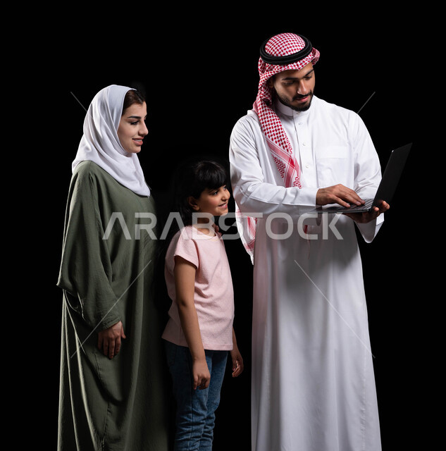 متابعة حصص الأونلاين وإجراء مكالمة فيديو من خلال الحاسوب المحمول، بورتريه لرجل عربي سعودي خليجي يحمل اللابتوب ويقف برفقة زوجته وابنته، الأجواء العائلية المليئة بالحب والترابط والإستقرار، مفهوم الإهتمام والإعتناء بالعائلة، خلفية سوداء