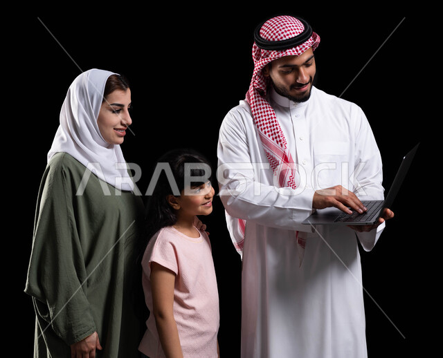 الأجواء العائلية المليئة بالحب والترابط والإستقرار، بورتريه لرجل عربي سعودي خليجي يحمل اللابتوب ويقف برفقة زوجته وابنته، مفهوم الإهتمام والإعتناء بالعائلة، متابعة حصص الأونلاين وإجراء مكالمة فيديو من خلال الحاسوب المحمول، خلفية سوداء