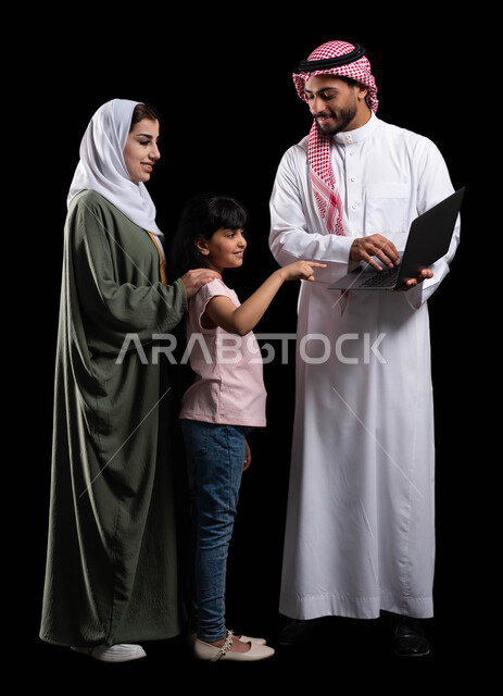 بورتريه لرجل عربي سعودي خليجي يحمل اللابتوب ويقف برفقة زوجته وابنته، الأجواء العائلية المليئة بالحب والترابط والإستقرار، متابعة حصص الأونلاين وإجراء مكالمة فيديو من خلال الحاسوب المحمول، صورة الجسم بالطول الكامل، خلفية سوداء