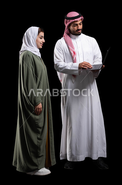 الحياة الزوجية الناجحة، التسوق الإلكتروني وتصفح الإنترنت من خلال الحاسوب المحمول، بورتريه لرجل عربي خليجي سعودي يستخدم اللابتوب برفقة زوجته، الترابط الأسري والمحافظة على أمان وإستقرار العائلة، صورة الجسم بالطول الكامل، خلفية سوداء