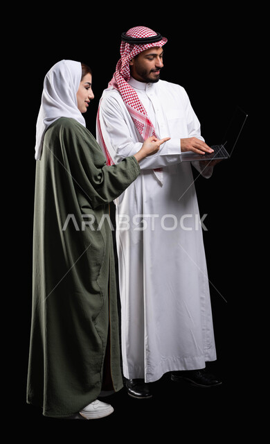 صورة الجسم بالطول الكامل, فريق عمل يقومون في التعاون والتشارك في إنجاز الأعمال, النظر و الإشارة إلى شيء ما في الحاسوب, مناقشة وتنظيم الاعمال, بورتريه لرجل أعمال عربي خليجي سعودية يحمل الحاسوب المحمول بجانبه موظفة عربية خليجية سعودية, خلفية سوداء