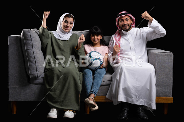 الشغف بمتابعة المباراة بإيماءات الانتصار والفرحة بالفوز, مفهوم الاهتمام والاعتناء بالأبناء, قضاء أوقات ممتعه بأجواء عائلية سعيده, بورتريه لعائلة عربية سعودية خليجية تقوم برفع يدها للأعلى بحماس, الجلوس في غرفة المعيشة والاستمتاع بتحقيق الأهداف, خلفية سوداء