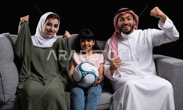 مفهوم الاهتمام والاعتناء بالأبناء, الشغف بمتابعة المباراة بإيماءات الانتصار والفرحة بالفوز, قضاء أوقات ممتعه بأجواء عائلية سعيده, بورتريه لعائلة عربية سعودية خليجية تقوم برفع يدها للأعلى بحماس, الجلوس في غرفة المعيشة والاستمتاع بتحقيق الأهداف, خلفية سوداء