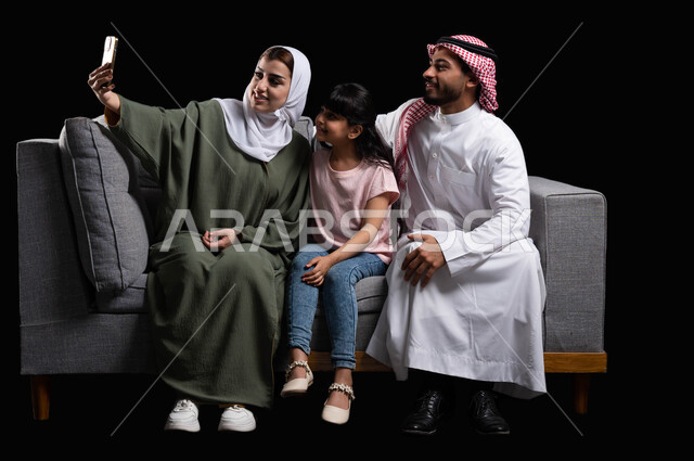 فرحة الأبناء بقضاء وقت ممتع مع الأهل, التقاط سيلفي عائلي تذكاري عبر الهاتف المحمول, بورتريه لعائلة عربية سعودية خليجية مبتسمة تقوم بتوثيق اللحظات الممتعة بإيماءات السعادة والفرح, الاجتماع الأسري في غرفة المعيشة وبناء مجتمع سعودي متنامي, خلفية سوداء