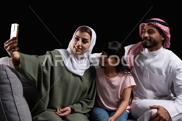 التقاط سيلفي عائلي تذكاري عبر الهاتف المحمول, بورتريه مقرب لعائلة عربية سعودية خليجية مبتسمة تقوم بتوثيق اللحظات الممتعة بإيماءات السعادة والفرح, الاجتماع الأسري في غرفة المعيشة وبناء مجتمع سعودي متنامي, فرحة الأبناء بقضاء وقت ممتع مع الأهل, خلفية سوداء