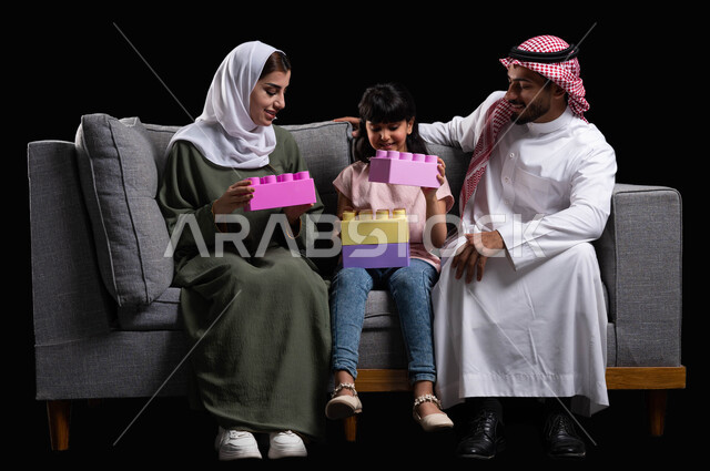 تعزيز الابتكارات الفكرية والذهنية للأبناء وقضاء وقت ممتع باللعب مع الأهل, الترابط الأسري والحفاظ على الاستقرار العائلي, بورتريه لعائلة عربية سعودية خليجية تجلس على الأريكة في غرفة المعيشة تقوم بنشاط تركيب المكعبات, رعاية وتشجيع مهارات الأولاد, خلفية سوداء