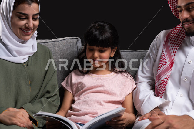 أهمية دور العائلة في تنمية ميول الأبناء, الأجواء العائلية المليئة بالحب والترابط والاستقرار, بورتريه مقرب لأسرة عربية خليجية سعودية تقوم بمساعدة ابنتهم على التعلم في المنزل, الجلوس على الكنبة في غرفة المعيشة وقضاء وقت ممتع في قراءة القصص, خلفية سوداء