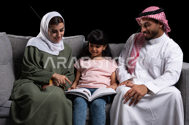 الجلوس على الكنبة في غرفة المعيشة وقضاء وقت ممتع في قراءة القصص المفيدة, الأجواء العائلية المليئة بالحب والترابط والاستقرار, دور العائلة في تنمية ميول الأبناء, بورتريه لأسرة عربية خليجية سعودية تقوم بمساعدة ابنتهم على التعلم في المنزل, خلفية سوداء