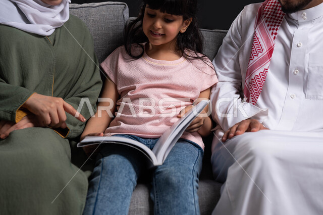 دور العائلة في تنمية ميول الأبناء, الأجواء العائلية المليئة بالحب والترابط والاستقرار, بورتريه مقرب لأسرة عربية خليجية سعودية تقوم بمساعدة ابنتهم على التعلم في المنزل, الجلوس على الكنبة في غرفة المعيشة وقضاء وقت ممتع في قراءة القصص المفيدة, خلفية سوداء