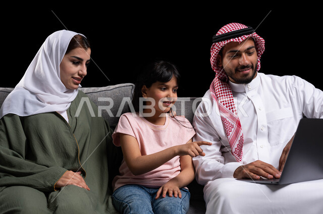 التفاعل مع العالم الافتراضي عن طريق مشاهدة الأفلام والمسلسلات والتسوق الالكتروني، إجراء مكالمات الفيديو والبثوث المباشره من خلال مواقع التواصل الاجتماعي، بورتريه مقرب لعائله عربيه خليجيه سعوديه تجلس على الأريكه أمام الحاسوب المحمول، خلفيه سوداء.