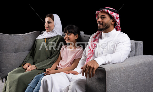 قضاء أوقات ممتعه وإنشاء روابط قويه مع الأبناء، بورتريه لأسرة عربية خليجية سعودية تجلس على أريكه رماديه وتبتسم تقوم بالنظر إلى شيء ما، ايماءات وجه تدل على السعاده والسرور، أجواء عائليه ترفيهية سعيده برفقه الأهل، خلفيه سوداء.