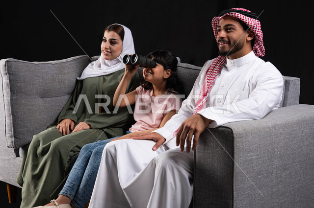 الاستمتاع والسرور بالجلسات العائليه الهادئه، دعم مواهب الأطفال وتنميه قدراتهم على حب الإستكشاف والتطلع للمستقبل، بورتريه لعائله عربيه خليجيه سعوديه تجلس على الأريكه، طفله سعوديه تحمل المنظار وتقوم بالنظر إلى شيء ما، خلفيه سوداء.